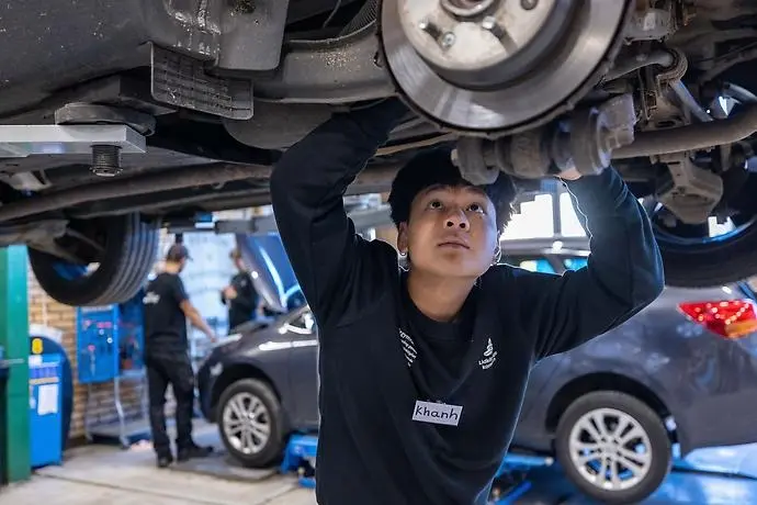 Lärare och elev i bilverkstad, undersöker upphissad personbil.