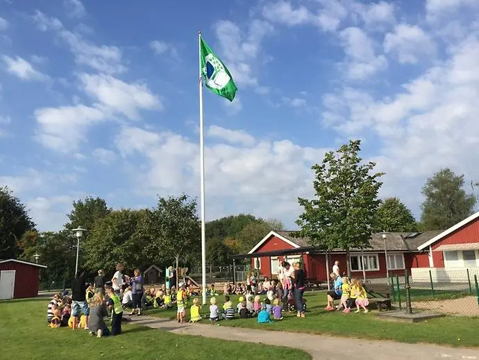 Majåkers röda träbyggnad och på gräset under flaggan sitter barn